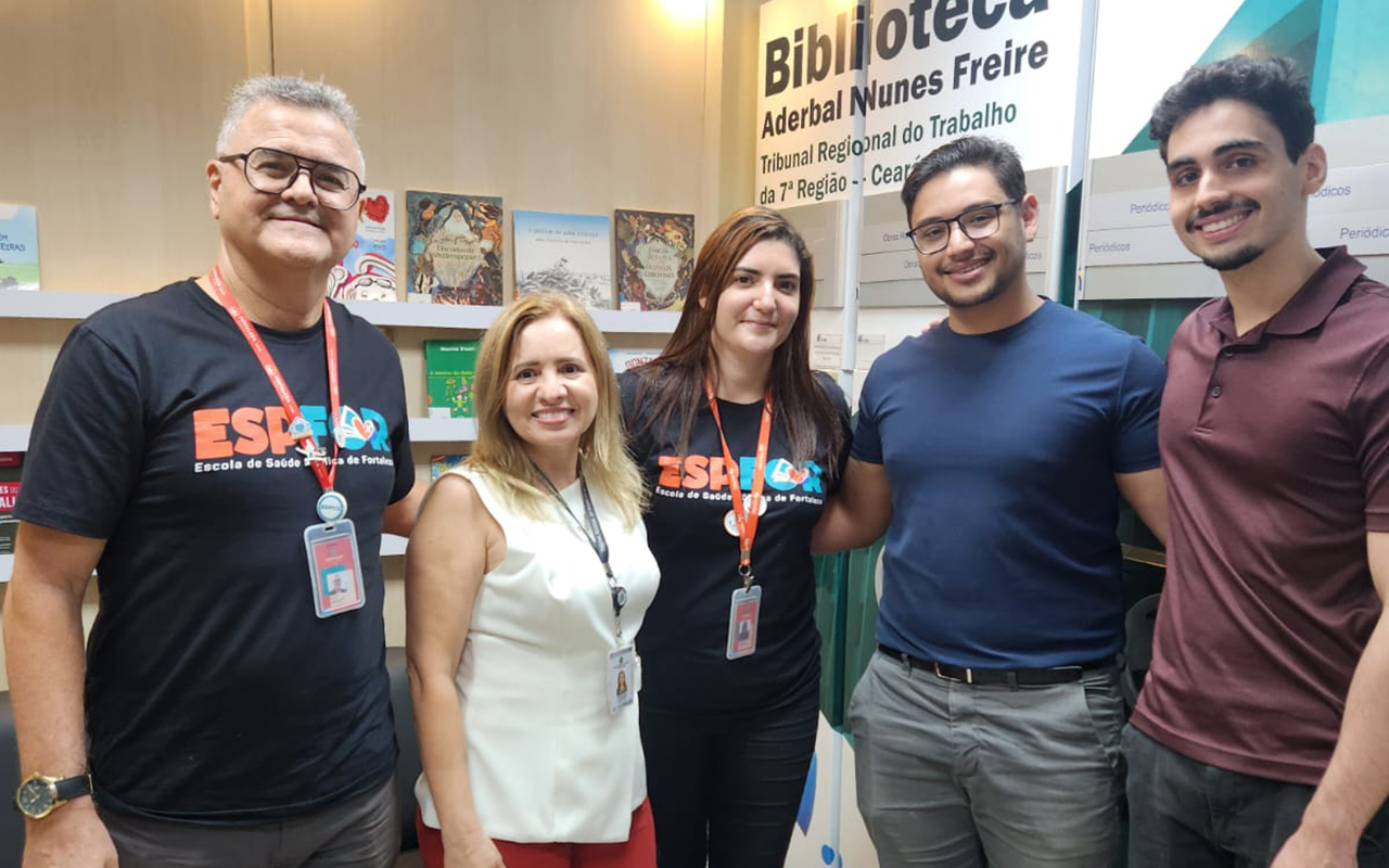 Fotografia colorida de um grupo de cinco pessoas em pé, posando sorrindo em um ambiente interno de biblioteca. Ao fundo, há estantes com livros expostos e um grande painel azul com o texto "Biblioteca Aderbal Nunes Freire, Tribunal Regional do Trabalho da 7ª Região - Ceará". Na frente, da esquerda para a direita, estão: um homem de camisa preta, uma mulher de blusa branca, uma mulher de camisa preta (ambas as mulheres e o primeiro homem usam crachás com a sigla ESFOP), um homem de camisa azul e um homem de camisa marrom. Eles estão próximos uns dos outros, com boa iluminação frontal.