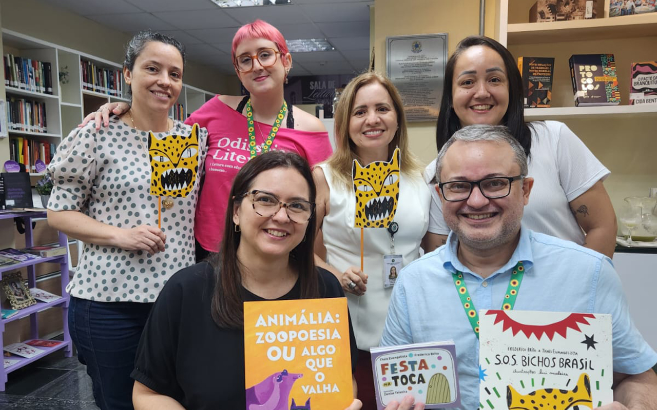Seis pessoas sorriem em uma biblioteca, com duas sentadas à frente de uma mesa redonda coberta por uma toalha verde com livros expostos. A mulher à esquerda segura o livro "Animália" e o homem à direita exibe "S.O.S. Bichos Brasil", enquanto as quatro mulheres em pé ao fundo seguram máscaras de papel com ilustrações de onças. O ambiente possui estantes brancas com livros e uma placa no teto com os dizeres "Convivência Literária".