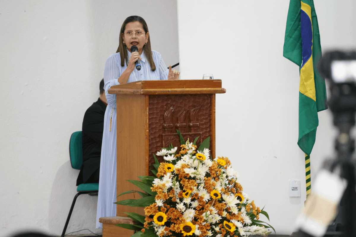 Card horizontal com fundo azul-marinho e grafismos geométricos em amarelo e azul claro. À esquerda, em uma moldura circular, a foto em preto e branco da Desembargadora Adriana Goulart de Sena Orsini, sorridente e vestindo blazer. À direita, o texto em branco e amarelo destaca seu nome e o título da palestra: "Trabalho Decente na Execução", com logomarcas institucionais nas bordas superior e inferior.