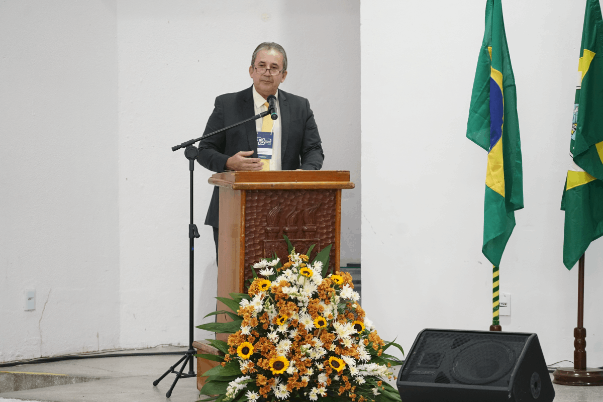 Card horizontal com fundo azul-marinho e grafismos em tons de amarelo e azul claro. À esquerda, a foto em preto e branco do Desembargador Paulo Régis Machado Botelho, que aparece sorrindo e usando terno. À direita, o texto destaca o nome do magistrado e o título da palestra: "A Justiça do Trabalho como Instituição de Equilíbrio Social", com as logomarcas institucionais da Justiça do Trabalho no topo e rodapé.