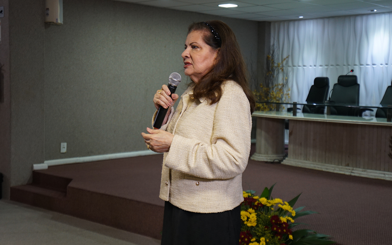 Fotografia em plano médio de uma mulher branca, de cabelos castanhos presos por uma tiara, falando ao microfone. Ela veste um casaco estruturado bege claro sobre uma blusa escura e está posicionada de perfil à esquerda da imagem. Ao fundo, vê-se a lateral de um palco acarpetado com um arranjo de flores coloridas e a bancada de autoridades vazia.