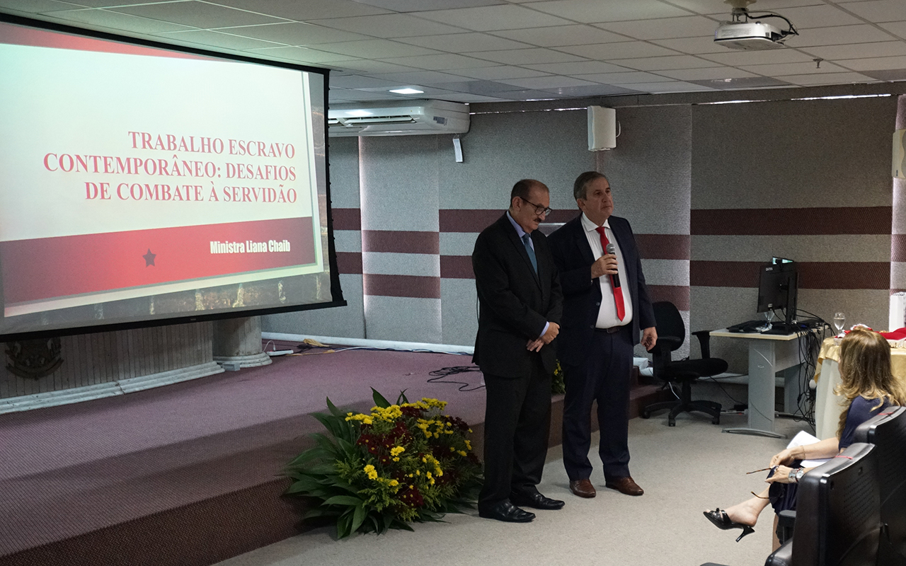 Fotografia em plano médio mostrando dois homens de terno e gravata em pé sobre um tablado de carpete bordô. À esquerda, uma tela de projeção exibe o título da palestra da ministra Liana Chaib. No canto inferior direito, uma mulher sentada de costas observa a apresentação, enquanto um arranjo de flores amarelas e vermelhas decora a base do palco.