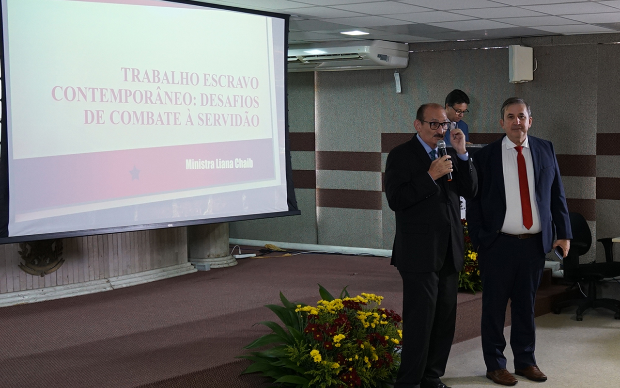 Fotografia em plano médio de dois homens de terno sobre um palco acarpetado. O homem à esquerda, de óculos e terno preto, fala ao microfone enquanto o outro, de terno azul e gravata vermelha, observa ao seu lado. À esquerda da dupla, uma grande tela branca exibe em letras vermelhas o título da palestra da ministra Liana Chaib.