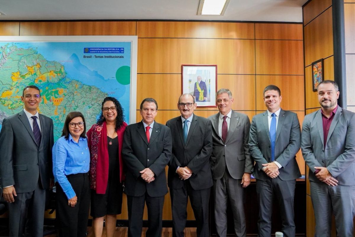 Seis homens e duas mulheres posam lado a lado em um gabinete oficial com paredes de madeira. A maioria veste trajes formais, como ternos e camisas sociais em tons de cinza, preto e azul. Ao fundo, à esquerda, destaca-se um grande mapa do Brasil com o título "Temas Institucionais" e, ao centro, uma foto emoldurada do Presidente da República. Todos sorriem para a câmera, transmitindo um clima de cooperação institucional.