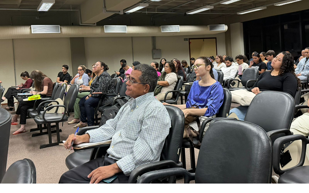 Uma plateia diversificada, composta por jovens e adultos sentados em poltronas de auditório pretas, assiste atentamente a um evento. No primeiro plano, um homem de camisa quadriculada e óculos faz anotações com uma caneta azul, ladeado por uma jovem de blusa azul cobalto. Ao fundo, as filas de assentos estão repletas de pessoas, algumas prestando atenção, outras olhando para os lados, sob a iluminação de luminárias fluorescentes no teto.