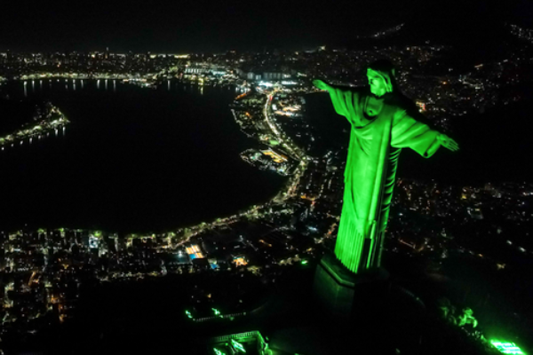 Vista aérea noturna do Cristo Redentor, no Rio de Janeiro, iluminado por uma vibrante luz verde. A estátua está em primeiro plano à direita, de braços abertos sobre a cidade sombria. Ao fundo, as luzes douradas da urbanização contornam a Lagoa Rodrigo de Freitas e a orla marítima, criando um forte contraste com o céu e as águas escuras. A perspectiva de cima ressalta a grandiosidade do monumento e a geografia da costa carioca.