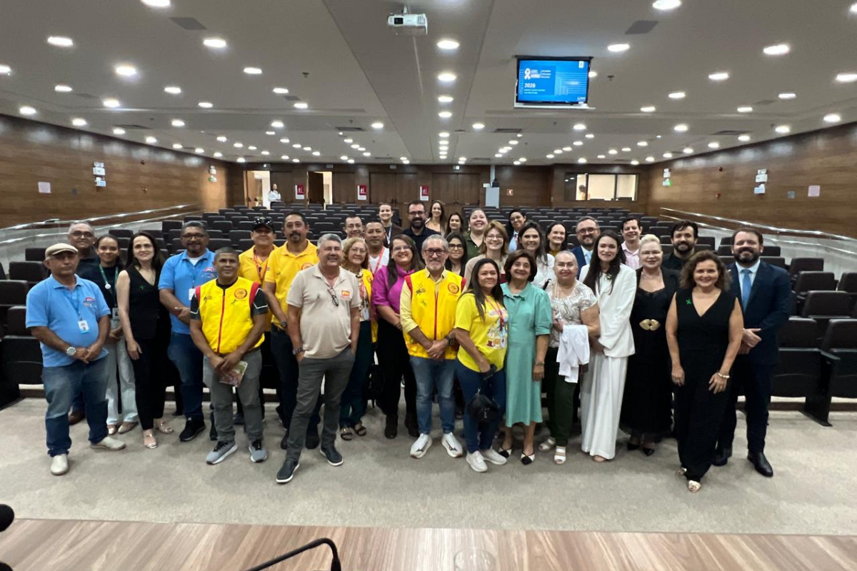 Em um auditório amplo com poltronas escuras ao fundo, um grupo diversificado de cerca de 30 pessoas posa sorridente para a foto. Alguns vestem trajes sociais e outros usam uniformes casuais, como camisas polo azuis e coletes amarelos. O ambiente é bem iluminado por várias luzes embutidas no teto, onde também se vê um projetor e um monitor exibindo a campanha "Abril Verde". A imagem transmite um clima de encerramento de evento coletivo e integração.