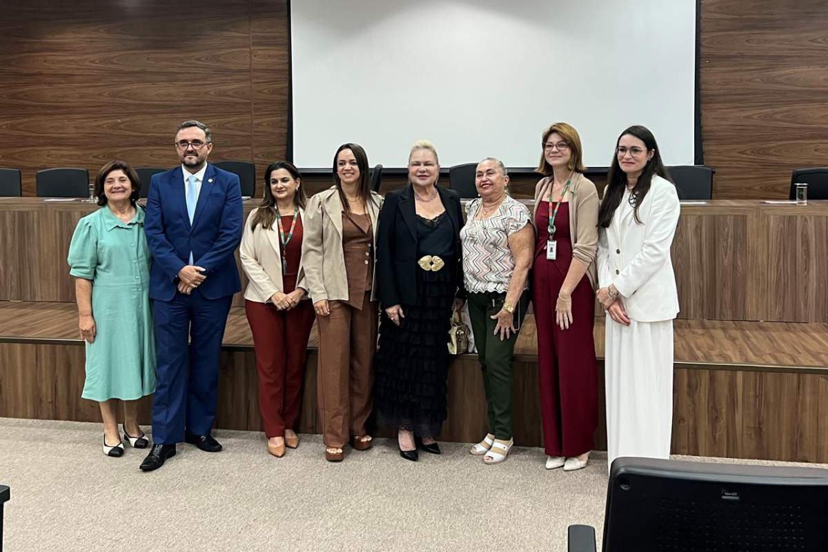 Em um auditório com painéis de madeira ao fundo, um grupo de oito pessoas — sete mulheres e um homem — posa em pé para uma foto oficial. Eles estão alinhados à frente de uma bancada elevada, vestindo trajes sociais em tons de verde, azul, marrom, preto, vermelho e branco. Ao fundo, uma tela de projeção branca está centralizada acima da bancada, completando o cenário formal e iluminado.