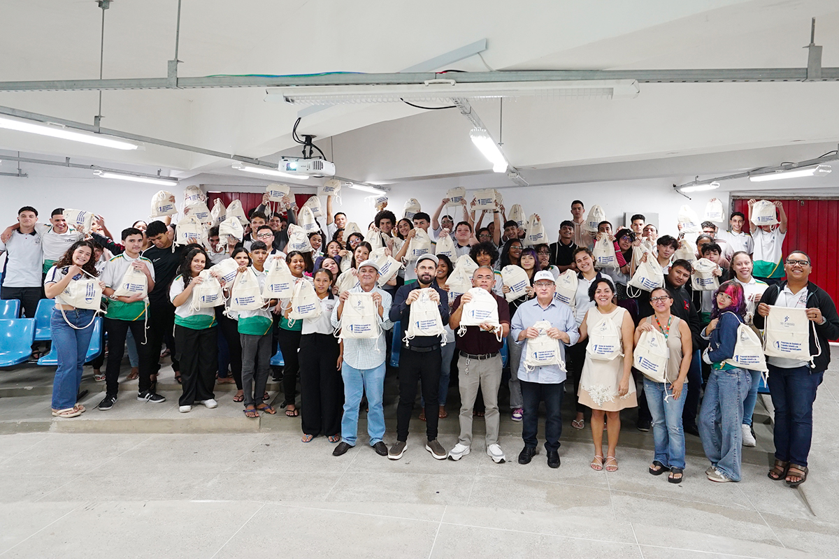 Uma foto em grupo num amplo salão mostra dezenas de jovens e alguns adultos em pé, sorrindo e segurando mochilas de pano idênticas para o alto. Os jovens vestem uniformes escolares brancos e verdes. À frente, os coordenadores e autoridades alinham-se ao centro, reforçando o clima de celebração e união do evento educativo.