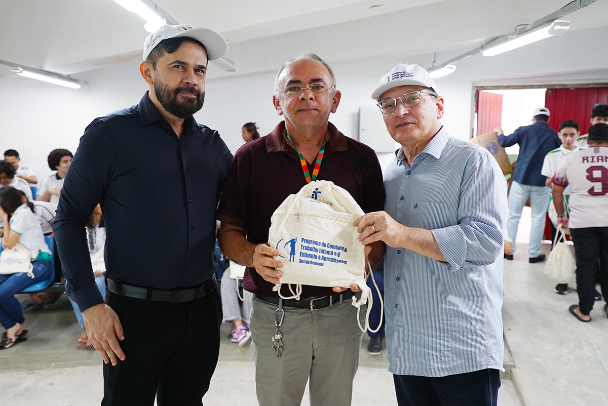 Três homens adultos posam lado a lado em plano médio, segurando juntos uma mochila tipo saco de cor crua. O saco exibe o logotipo do "Programa de Combate ao Trabalho Infantil". Eles vestem camisas casuais e dois usam bonés brancos. Ao fundo, jovens sentados em cadeiras azuis observam a cena em um ambiente iluminado.