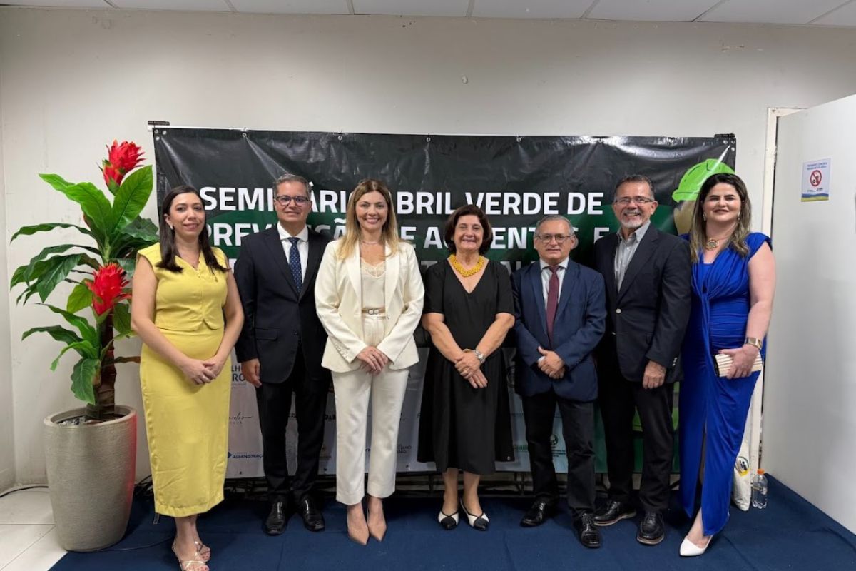 Sete pessoas (quatro mulheres e três homens) posam lado a lado em um evento formal. Ao fundo, um banner preto anuncia o Seminário Abril Verde, focado na prevenção de acidentes de trabalho. O grupo veste trajes sociais em cores como amarelo, branco, preto e azul royal, posicionados sobre um tapete azul. À esquerda, destaca-se um vaso com flores vermelhas, e à direita, uma porta branca completa o cenário.