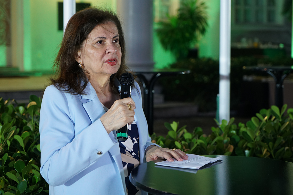 Fotografia em plano médio mostra uma mulher de pele clara e cabelos castanhos na altura dos ombros falando ao microfone. Ela veste um blazer azul-claro sobre uma blusa estampada, tem a mão esquerda apoiada sobre papéis em uma mesa alta e direciona o olhar para a direita. Ao fundo, arbustos verdes e uma estrutura arquitetônica iluminada completam o cenário noturno.