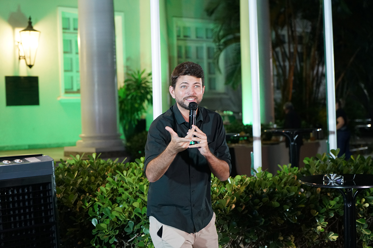 Fotografia em plano médio mostra um homem de pele clara, cabelos e barba castanhos curtos, falando ao microfone em um evento noturno. Ele veste camisa de botão preta e calça cáqui, sorri e gesticula com a mão direita aberta enquanto faz sua apresentação. Ao fundo, destacam-se arbustos verdes, colunas brancas e a fachada de um prédio histórico iluminada com luz verde.
