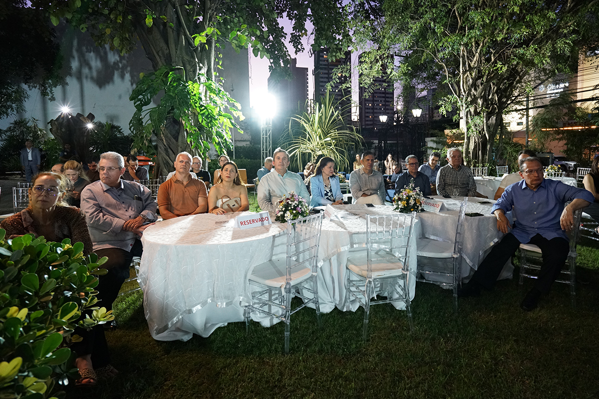 Fotografia em plano aberto mostra um grupo de pessoas sentadas ao redor de mesas redondas com toalhas brancas e arranjos de flores, dispostas sobre o gramado de um jardim. Os convidados estão voltados para a esquerda, prestando atenção ao evento, e sobre as mesas há pequenas placas sinalizadoras com a palavra "Reservado". Ao fundo, árvores frondosas, postes de iluminação e prédios urbanos compõem o cenário ao anoitecer.
