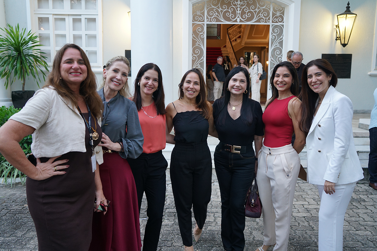 Fotografia em plano médio mostra sete mulheres sorridentes posando enfileiradas lado a lado em um piso de blocos intertravados. Elas usam trajes sociais em tons de marrom, vermelho, preto e branco, destacando-se uma delas à direita de terno totalmente branco. Ao fundo, vê-se a entrada de um prédio histórico de fachada clara, com uma imponente porta de ferro trabalhado aberta e uma escadaria interna ao fundo.