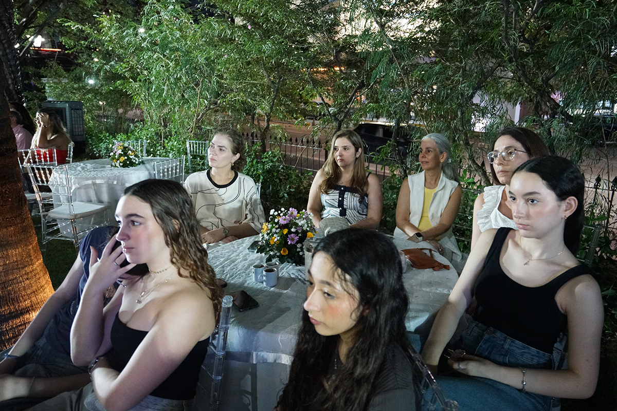otografia em plano aberto mostra um grupo de mulheres de diferentes idades sentadas ao redor de uma mesa redonda em um evento noturno ao ar livre. Elas estão posicionadas em um jardim gramado, com folhagens verdes e uma grade de ferro ao fundo, e direcionam a atenção para o lado esquerdo. Em primeiro plano, destacam-se três jovens de cabelos escuros vestindo blusas de alças em tons escuros.