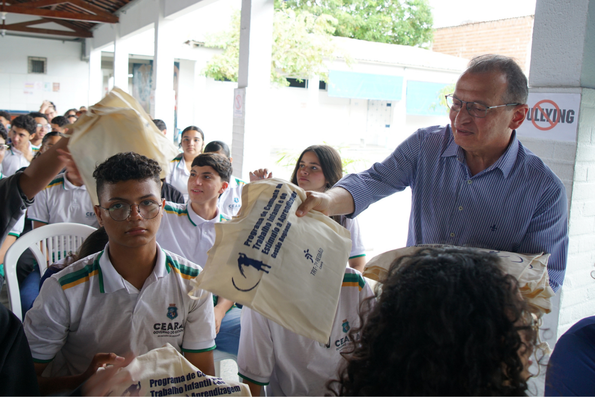Fotografia em plano médio de um homem de óculos e camisa listrada distribuindo sacolas ecológicas para estudantes sentados. À direita, ele sorri ao entregar uma sacola bege com inscrições em azul sobre o combate ao trabalho infantil. À esquerda, os jovens usam uniformes escolares brancos com detalhes em verde e amarelo. O evento ocorre em um pátio coberto com colunas brancas e árvores ao fundo.