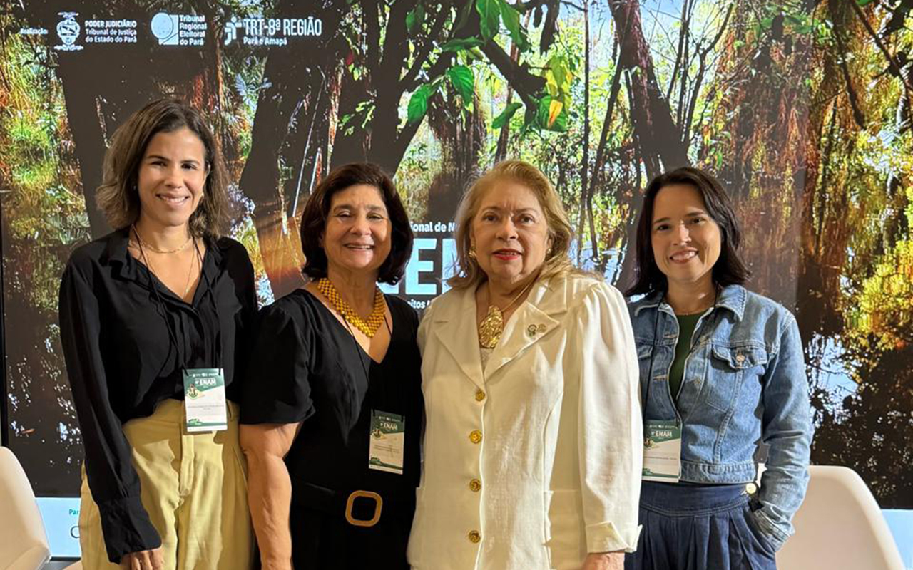 Quatro mulheres sorriem em pé, lado a lado, em cima de um palco com poltronas brancas ao fundo. Elas vestem trajes sociais e casuais finos em tons de preto, amarelo-claro, branco e jeans, usando crachás do evento. Atrás delas, um grande telão luminoso exibe uma imagem de floresta com logomarcas dos tribunais realizadores no topo.