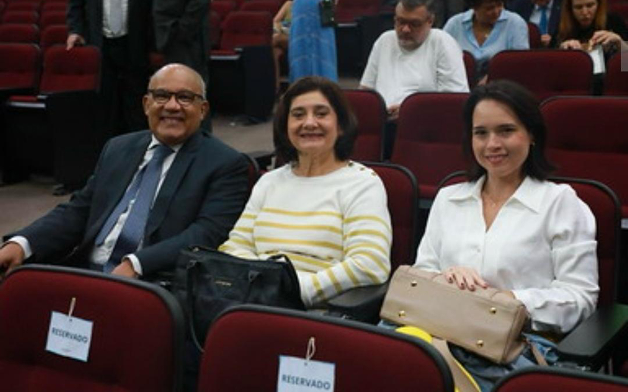Um homem de terno escuro e duas mulheres com blusas brancas — uma delas com listras amarelas — sorriem sentados em poltronas vermelhas de um auditório. À frente deles, as poltronas trazem placas com o aviso "Reservado" fixadas nos encostos. Ao fundo, outras pessoas se movimentam ou estão sentadas nas fileiras superiores do espaço.