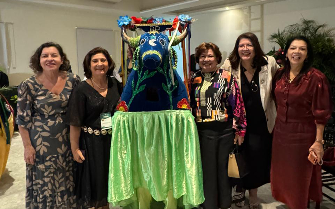 Cinco mulheres em trajes elegantes posam sorridentes em um salão iluminado, distribuídas ao redor de uma fantasia folclórica de boi-bumbá. A representação do boi é azul, decorada com flores e fitas coloridas, e possui uma saia de tecido verde acetinado que cobre as pernas do dançarino. Ao fundo, o ambiente apresenta paredes claras e detalhes de plantas ornamentais.