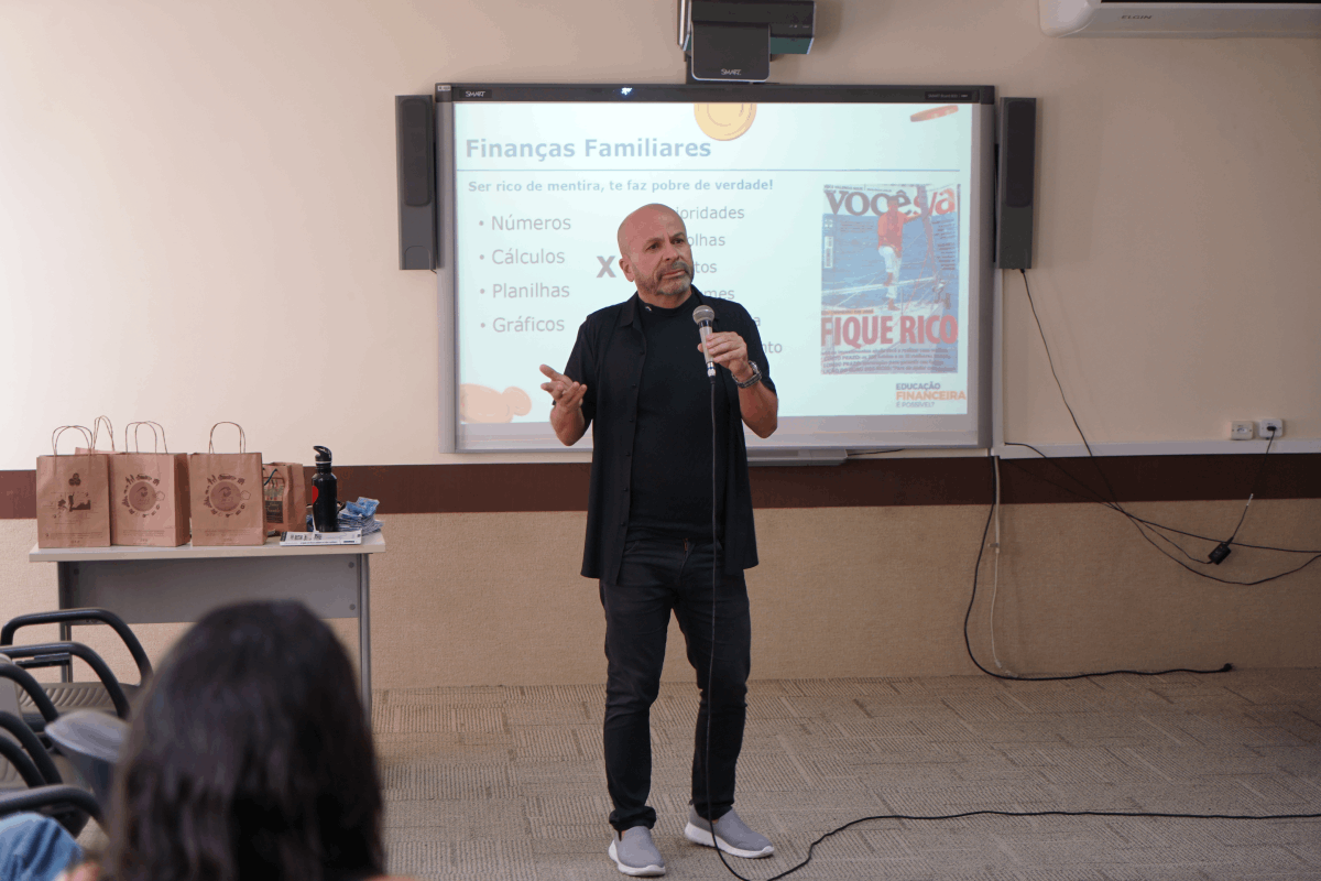 Um homem careca, vestindo camisa e calça pretas, fala ao microfone diante de uma tela de projeção com o título "Finanças Familiares". À esquerda, há uma mesa com sacolas de papel pardo, e em primeiro plano, vê-se a parte de trás da cabeça de uma espectadora. O ambiente é uma sala de aula ou auditório com piso cinza e paredes claras.
