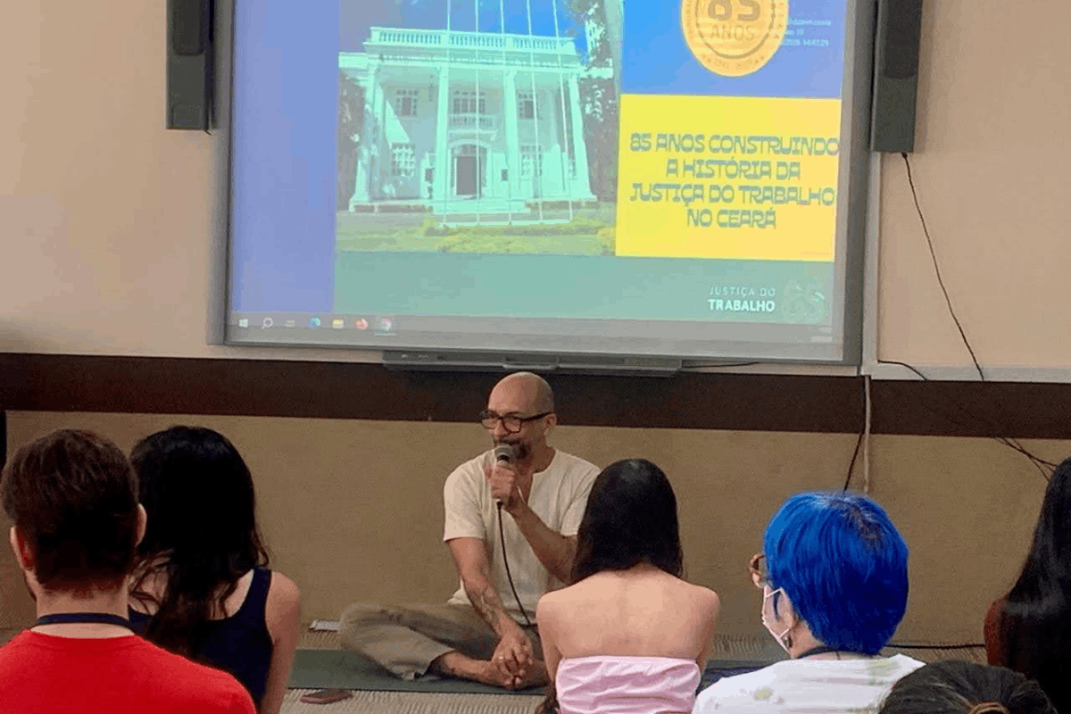 Um homem de óculos e camiseta clara fala ao microfone, sentado de pernas cruzadas no chão em frente a uma plateia. Atrás dele, uma grande tela de projeção exibe a imagem de um prédio histórico com o texto "85 anos construindo a história da Justiça do Trabalho no Ceará". O público, visto de costas, acompanha a apresentação em um ambiente de palestra ou vivência.