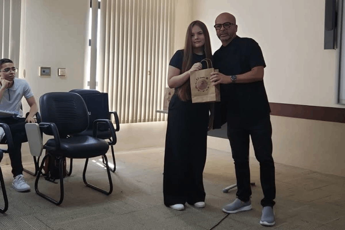Uma jovem de cabelos longos e lisos e um homem careca, de óculos, ambos vestindo roupas pretas, posam juntos de pé em uma sala. Eles seguram, entre si, uma sacola de papel pardo com uma estampa circular no centro. Ao fundo, uma janela com persianas verticais ilumina parcialmente o ambiente, que conta com cadeiras de escritório à esquerda.