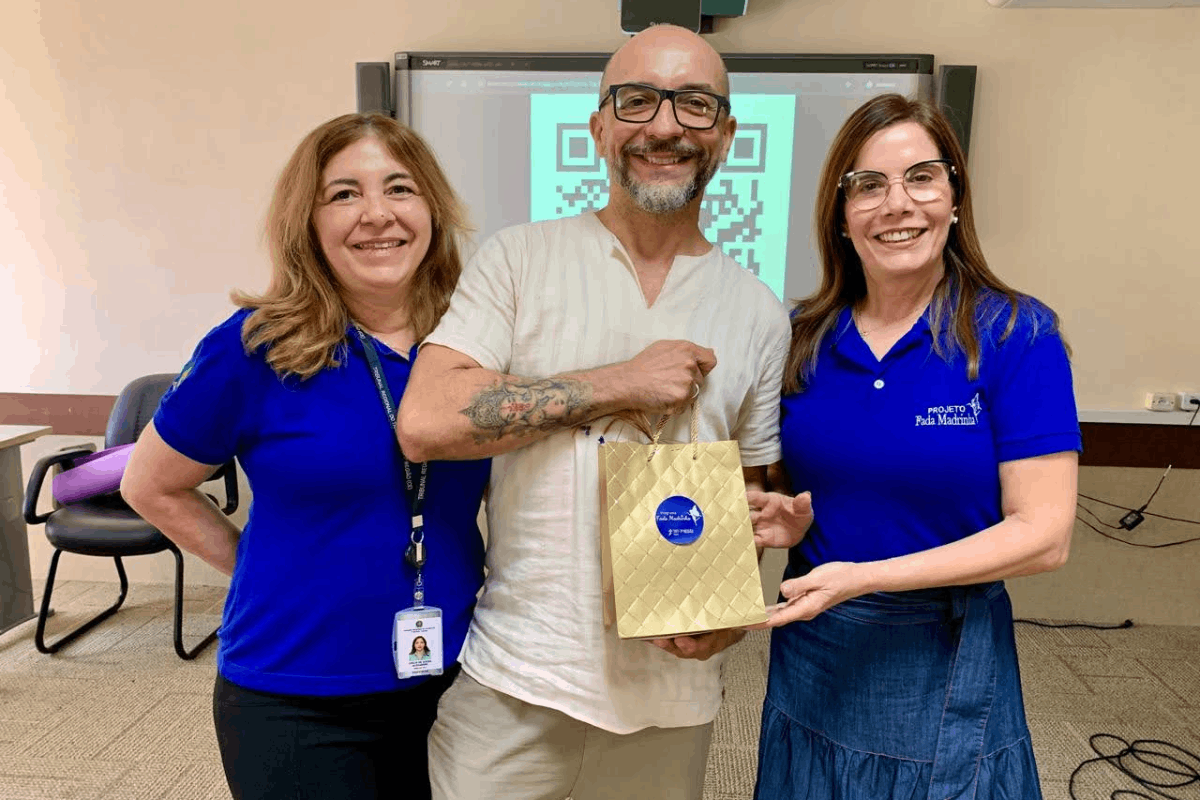 O homem sorridente de óculos e bata clara posa no centro, ladeado por duas mulheres vestindo camisas polo azuis, uma delas com a estampa "Projeto Fada Madrinha". Juntos, eles seguram uma sacola de presentes dourada com um adesivo redondo azul no meio. Ao fundo, uma tela digital exibe um QR Code em destaque no ambiente de sala de aula.