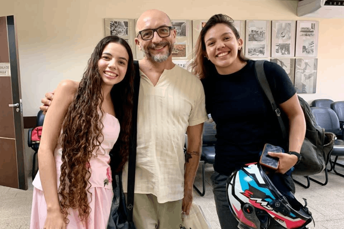 O homem de óculos e bata clara, agora sorridente e com os braços nos ombros das acompanhantes, posa entre duas jovens em uma sala de aula. À esquerda, uma jovem com longos cabelos cacheados usa vestido rosa, e à direita, outra jovem sorri vestindo camiseta preta e segurando um capacete colorido de motocicleta. Ao fundo, veem-se quadros com fotos na parede e cadeiras enfileiradas.