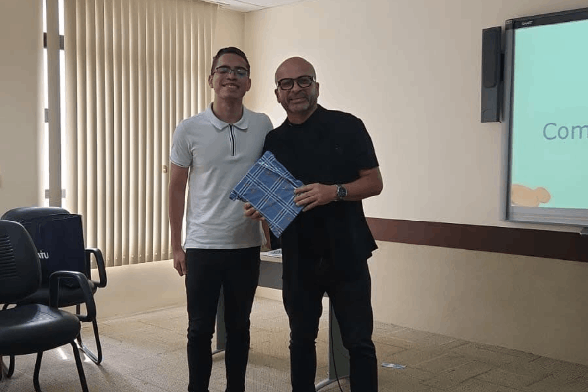 Um jovem sorridente de camisa polo branca e calça escura posa ao lado de um homem careca de óculos e roupa preta. Eles seguram juntos um embrulho azul com estampa xadrez em uma sala iluminada por uma grande janela com persianas verticais ao fundo. À direita, vê-se parte de uma tela de projeção e, à esquerda, cadeiras de escritório pretas.