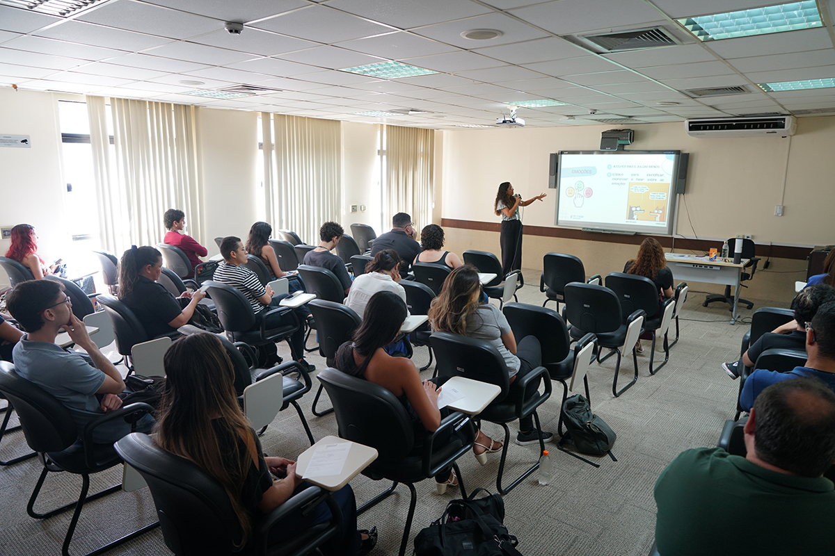 Foto ampla em perspectiva diagonal de uma sala de aula iluminada com estagiários sentados em cadeiras universitárias, assistindo a uma palestra. No fundo da sala, uma mulher de blusa estampada e calça escura está de pé, gesticulando em direção a uma tela digital que exibe um slide intitulado "EMOÇÕES". A luz natural entra pelas grandes janelas com persianas verticais ao lado esquerdo.
