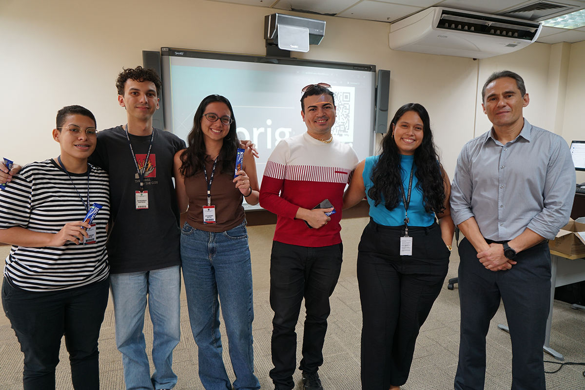 Foto em plano médio mostra um grupo de cinco jovens estagiários alinhados lado a lado com o palestrante Fernando Freitas, posicionado na extremidade direita. Eles estão sorrindo em um ambiente interno, e alguns dos estudantes seguram barras de chocolate azul como brindes, posicionados à frente de uma lousa digital que exibe a palavra "Obrigado".