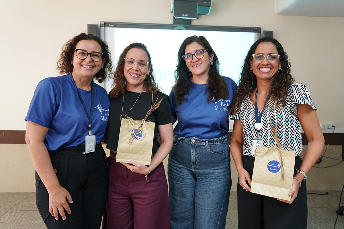 Quatro mulheres sorridentes estão posicionadas de pé, lado a lado, em frente a uma tela digital branca. Duas delas usam camisetas azuis escuras com o logotipo do "Programa Fada Madrinha" impresso, enquanto as outras duas seguram sacolas de presentes douradas com laços, em um momento de confraternização e encerramento.