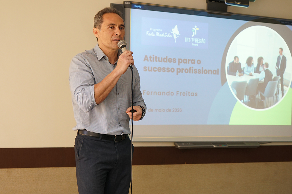 Imagem focada em Fernando Freitas, um homem de cabelos curtos grisalhos, vestindo camisa social cinza de mangas compridas dobradas e calça escura. Ele está de pé, segurando um microfone próximo à boca com a mão direita e um passador de slides na mão esquerda. Ao fundo, a tela digital exibe o título da palestra: "Atitudes para o sucesso profissional".