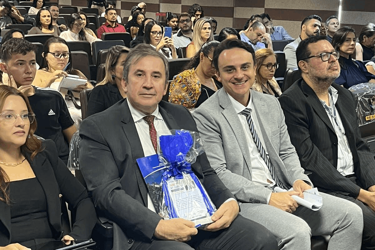 Em um auditório com público ao fundo, um homem de terno escuro e gravata vermelha está sentado em primeiro plano, segurando um presente embalado com laço azul. À sua esquerda, um homem sorridente de terno cinza e gravata listrada o acompanha, enquanto outras pessoas nas laterais e fileiras de trás observam o evento.