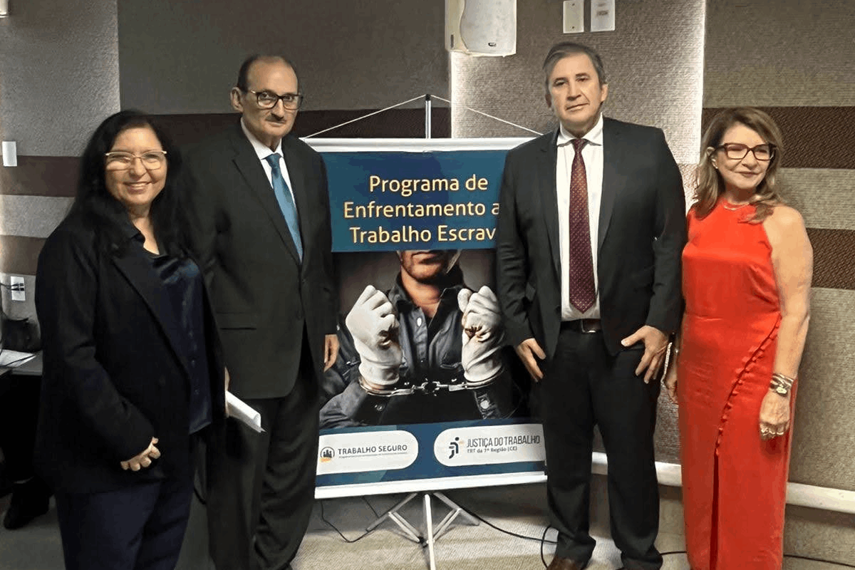 Quatro pessoas posam sorridentes, de pé, ao lado de um banner vertical sobre o combate ao trabalho escravo. À esquerda, uma mulher de blazer escuro e um homem de terno preto e gravata azul; à direita, um homem de terno preto com gravata vermelha e uma mulher de vestido longo vermelho. O banner centralizado mostra uma imagem em preto e branco de mãos algemadas e os logotipos da Justiça do Trabalho e Trabalho Seguro.