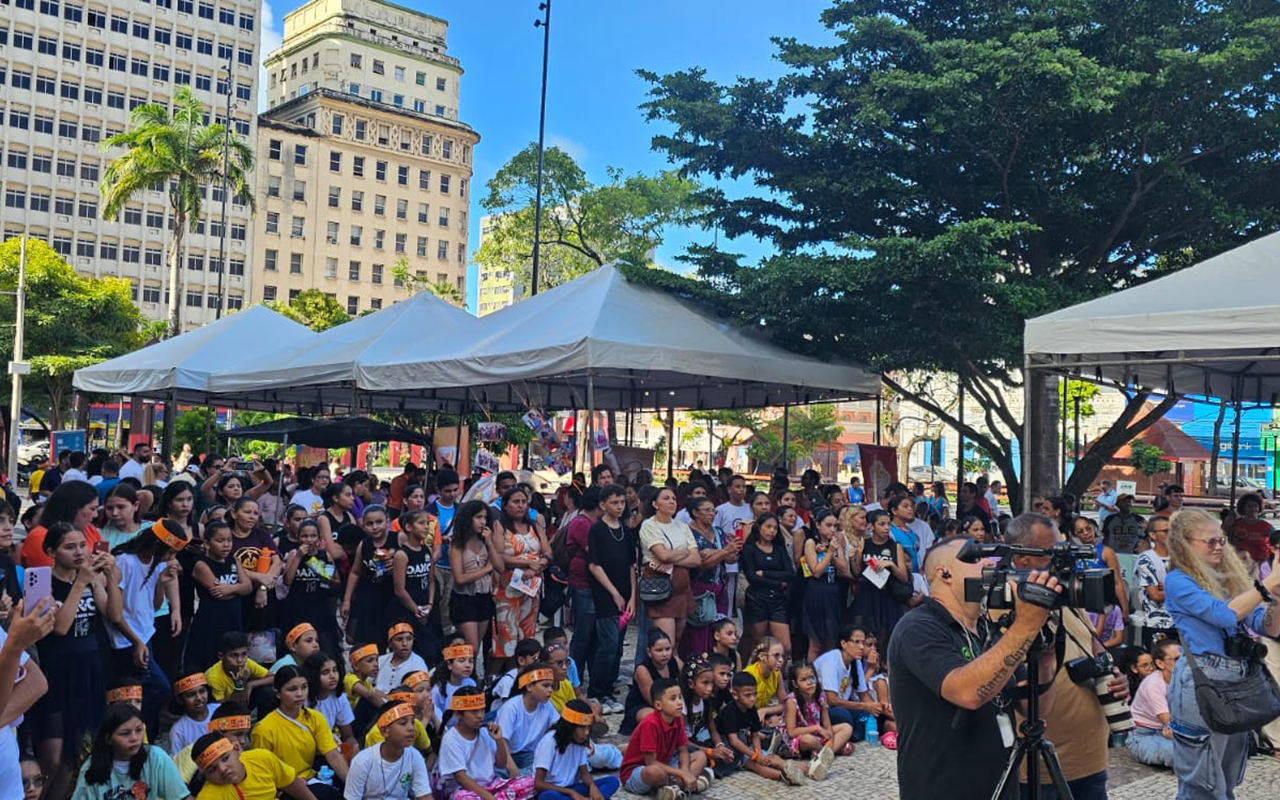 Uma foto panorâmica da Praça do Ferreira, em Fortaleza, mostra uma grande aglomeração de pessoas sentadas e em pé. No primeiro plano, à esquerda, diversas crianças e adolescentes vestem camisetas uniformizadas e faixas na cabeça, acompanhadas por adultos. No primeiro plano, à direita, um cinegrafista foca sua câmera profissional em um tripé na direção oposta, ladeado por outra pessoa tirando fotos com um smartphone. Ao fundo, sob árvores frondosas, diversas tendas brancas de eventos estão montadas, com prédios altos e antigos na paisagem urbana.