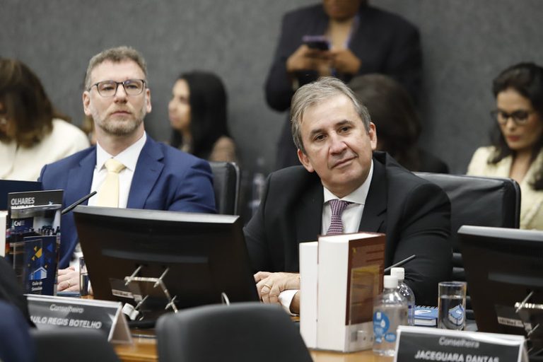 otografia em plano médio de um homem de terno azul e gravata amarela sentado à mesa de reuniões, olhando em direção à câmera. Ele segura um relatório aberto com gráficos, posicionado atrás de uma placa de identificação preta onde se lê "Conselheiro PAULO REGIS MACHADO BOTELHO - TRT-7". Ao fundo, em ambiente de escritório com detalhes em madeira, outras pessoas aparecem desfocadas em pé e sentadas