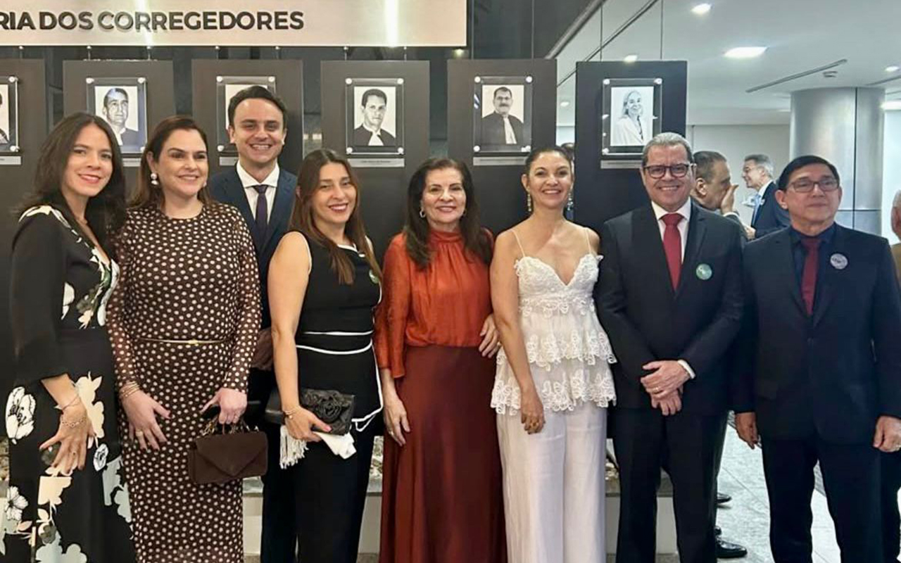 Fotografia em plano inteiro de um grupo de oito pessoas — cinco mulheres e três homens — sorrindo alinhadas lado a lado em um ambiente interno iluminado. Eles vestem trajes formais e posam à frente de uma parede preta de galeria onde se lê parcialmente "RIA DOS CORREGEDORES" acima de retratos em preto e branco emoldurados. Ao fundo, à direita, há uma coluna metálica e outras pessoas conversando dispersas pelo salão.