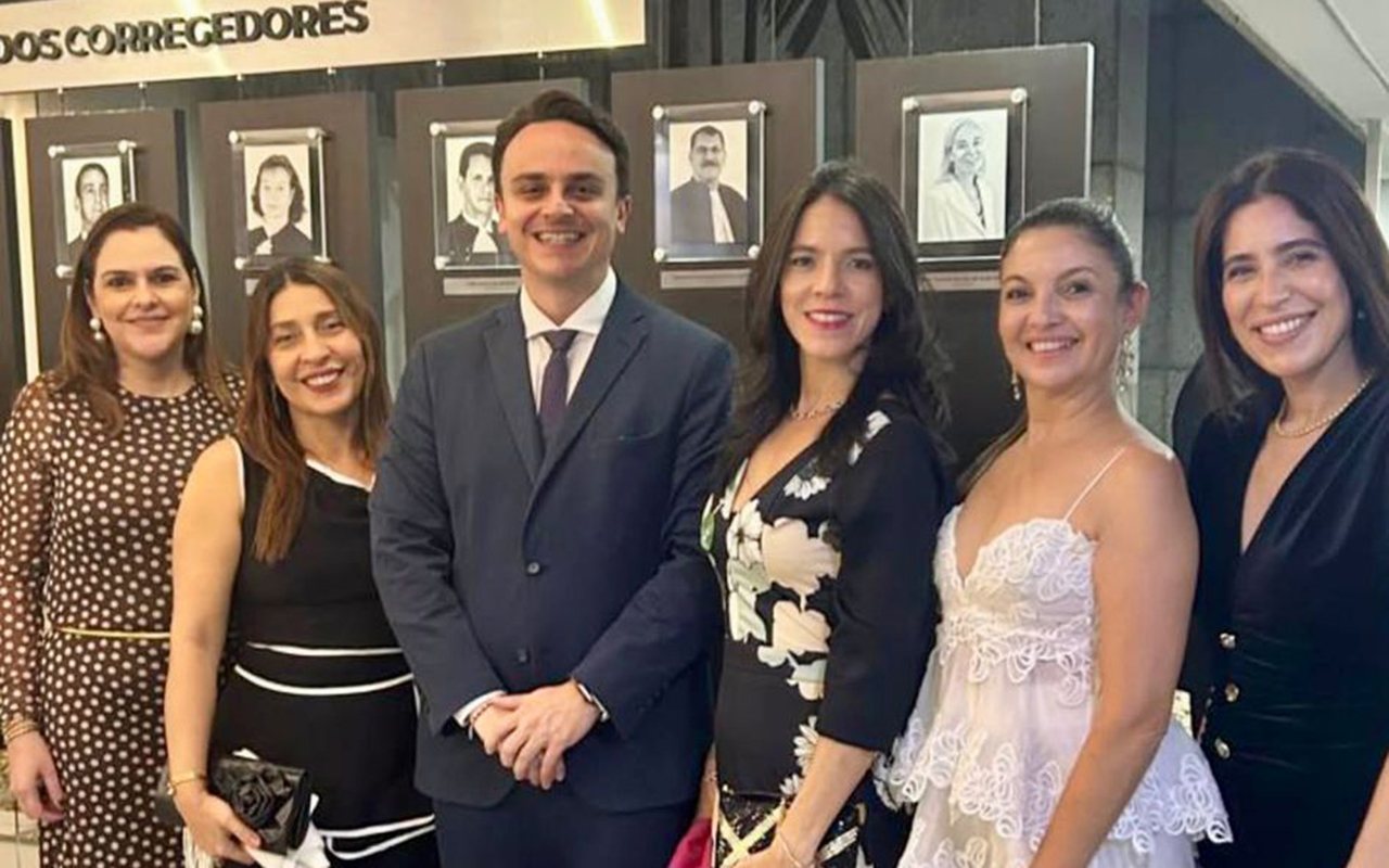 Fotografia em plano médio de seis pessoas — cinco mulheres e um homem — sorrindo alinhadas lado a lado em um evento formal. Da esquerda para a direita, a primeira mulher usa vestido marrom de poá; a segunda, vestido preto com detalhes brancos; o terceiro é um homem de terno azul-escuro; a quarta veste vestido preto floral; a quinta usa vestido branco rendado de alças; e a sexta, um vestido preto de decote transpassado. Ao fundo, há uma galeria de retratos emoldurados sob o letreiro parcial "DOS CORREGEDORES".