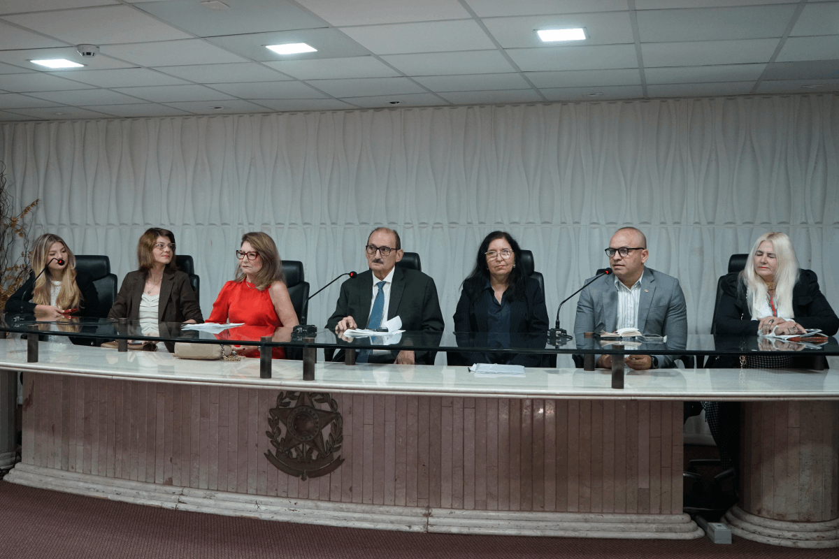 Sete pessoas estão sentadas lado a lado atrás de uma longa bancada curva de mármore claro, que exibe o brasão da República do Brasil na parte central inferior. O grupo, composto por cinco mulheres e dois homens com roupas formais, está posicionado em frente a uma parede branca com texturas onduladas verticais. Diante de cada participante há microfones de haste flexível e papéis espalhados sobre a superfície de vidro da mesa