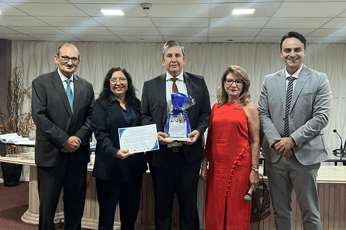 Uma mulher de cabelos longos e terninho escuro sorri no centro, ladeada por dois homens de terno e óculos, posicionados de pé em um auditório. Ao fundo, vê-se uma longa bancada de mármore claro com tampo de vidro, cadeiras executivas pretas e uma parede texturizada com ondulações verticais brancas. O chão é coberto por um carpete avermelhado e o ambiente é iluminado por luminárias embutidas no teto.