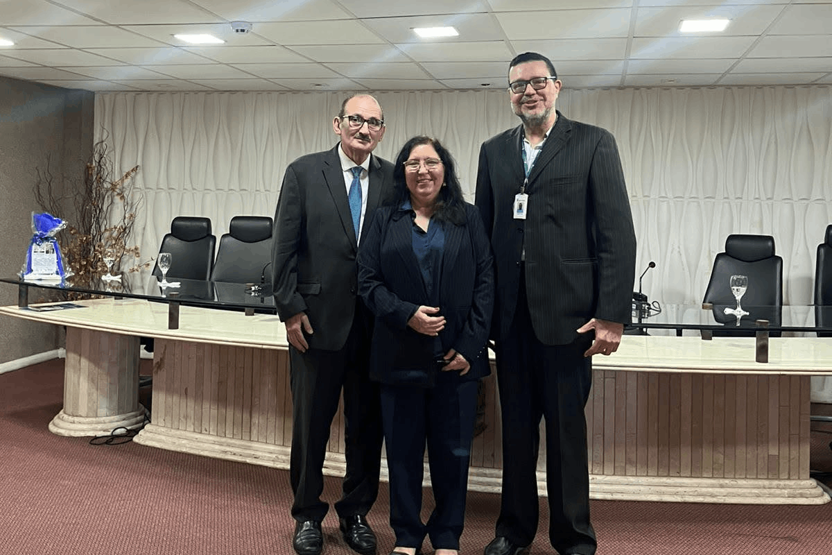 Uma mulher de cabelos longos e terninho escuro sorri no centro, ladeada por dois homens de terno e óculos, posicionados de pé em um auditório. Ao fundo, vê-se uma longa bancada de mármore claro com tampo de vidro, cadeiras executivas pretas e uma parede texturizada com ondulações verticais brancas. O chão é coberto por um carpete avermelhado e o ambiente é iluminado por luminárias embutidas no teto.