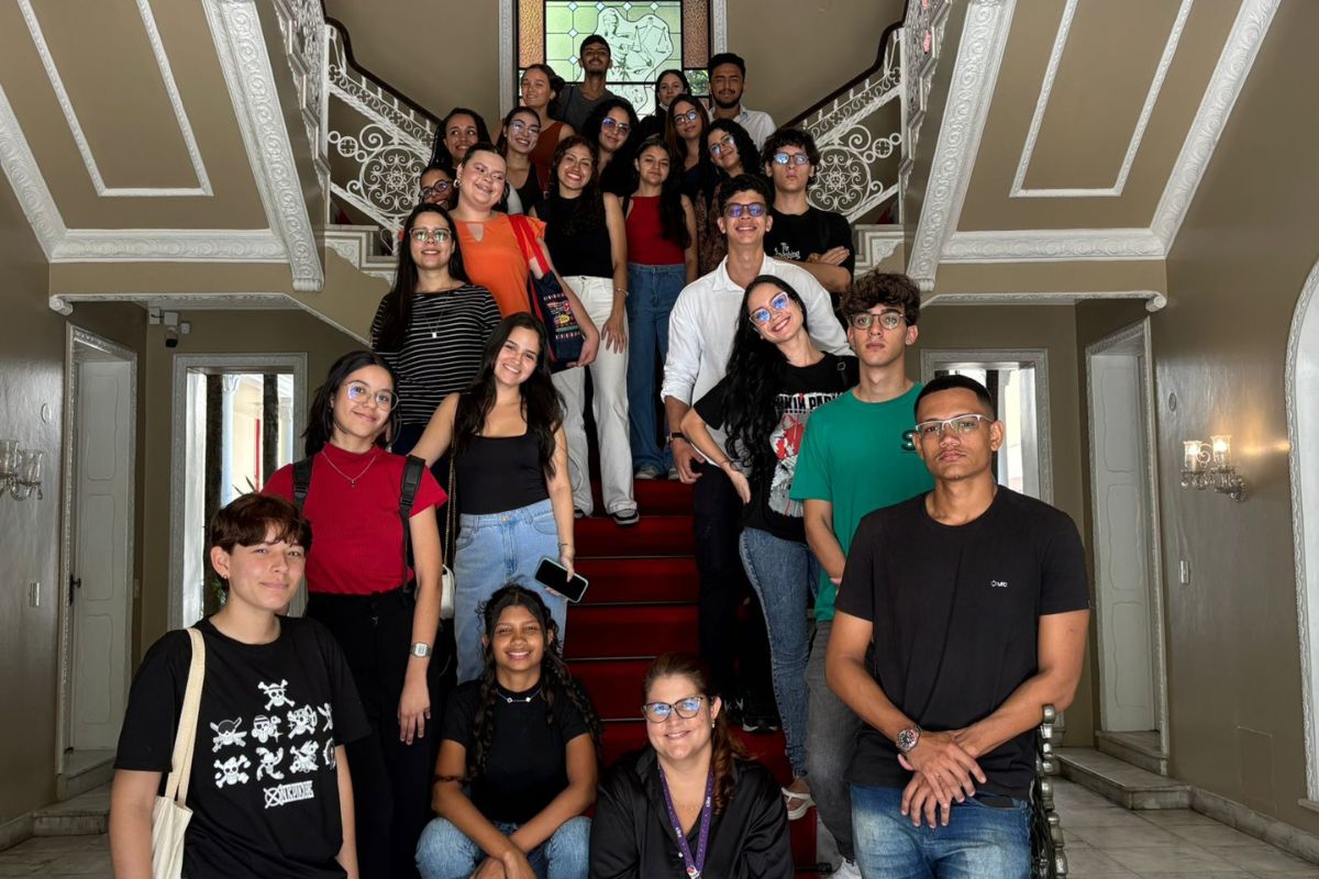 Fotografia de um grupo de cerca de vinte jovens e adultos sorridentes, posando dispostos ao longo de uma imponente escadaria interna com tapete vermelho e corrimão detalhado. À frente, algumas pessoas estão sentadas ou em pé no primeiro plano, enquanto as demais se distribuem pelos degraus acima, sob a luz de um ambiente clássico com paredes em tons pastéis.
