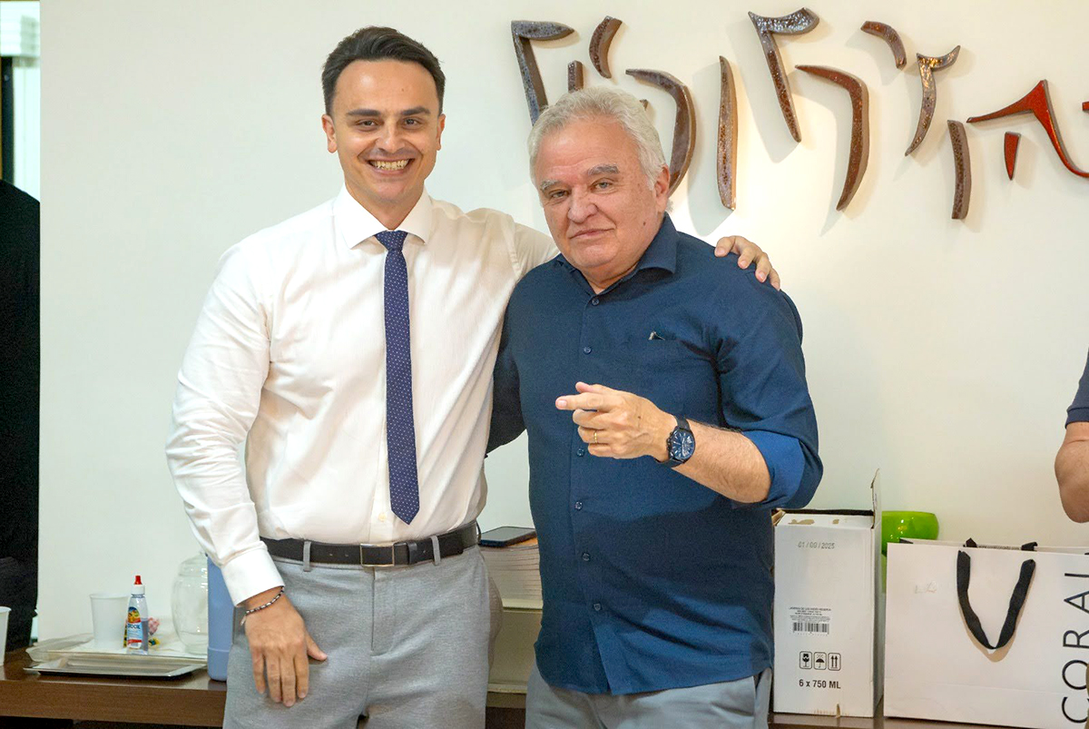 Fotografia colorida de dois homens em pé, abraçados pelos ombros e sorrindo. À esquerda, um homem jovem veste camisa branca, gravata azul e calça cinza; à direita, um homem de cabelos grisalhos usa camisa polo azul-marinho. Ao fundo, uma parede clara exibe uma escultura metálica de caracteres estilizados, sobre um balcão com caixas e objetos diversos.