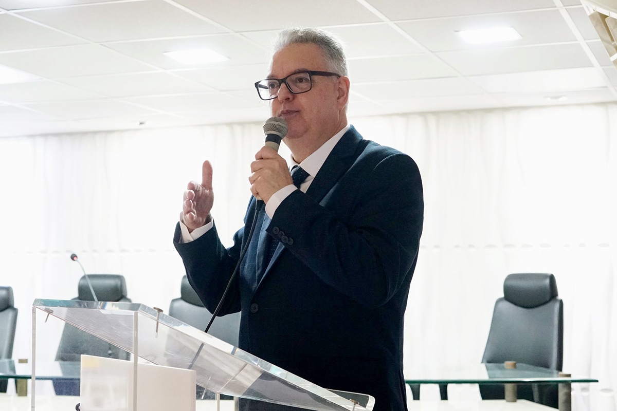 Plano médio do palestrante grisalho, vestindo terno azul-marinho e gravata, falando ao microfone. Ele está posicionado atrás de um púlpito de acrílico transparente, com a mão direita levemente levantada em gesto explicativo. O fundo é composto por cortinas brancas e cadeiras de escritório pretas sob luzes de teto embutidas.