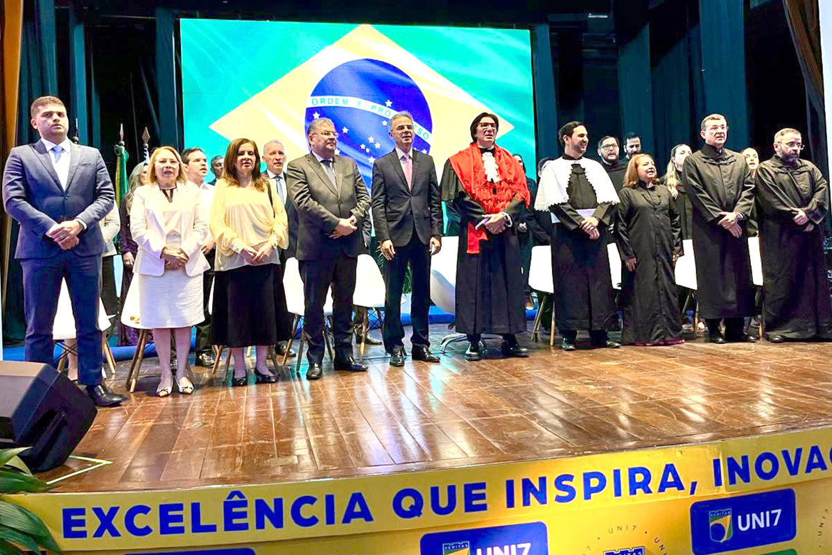 Plano geral de uma cerimônia em um palco com piso de madeira. Diversas autoridades e acadêmicos estão perfilados à frente de um grande telão que projeta a bandeira do Brasil. Na base do palco, lê-se em destaque a frase: "EXCELÊNCIA QUE INSPIRA, INOVAÇÃO...".