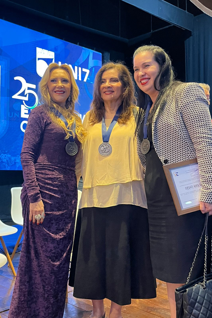 Três mulheres sorridentes posam juntas em primeiro plano. As duas à esquerda usam medalhas prateadas com o número "25" gravado; a da direita segura um certificado. Elas vestem, respectivamente, um vestido roxo aveludado, uma blusa bege com saia preta e um conjunto preto com blazer estampado.