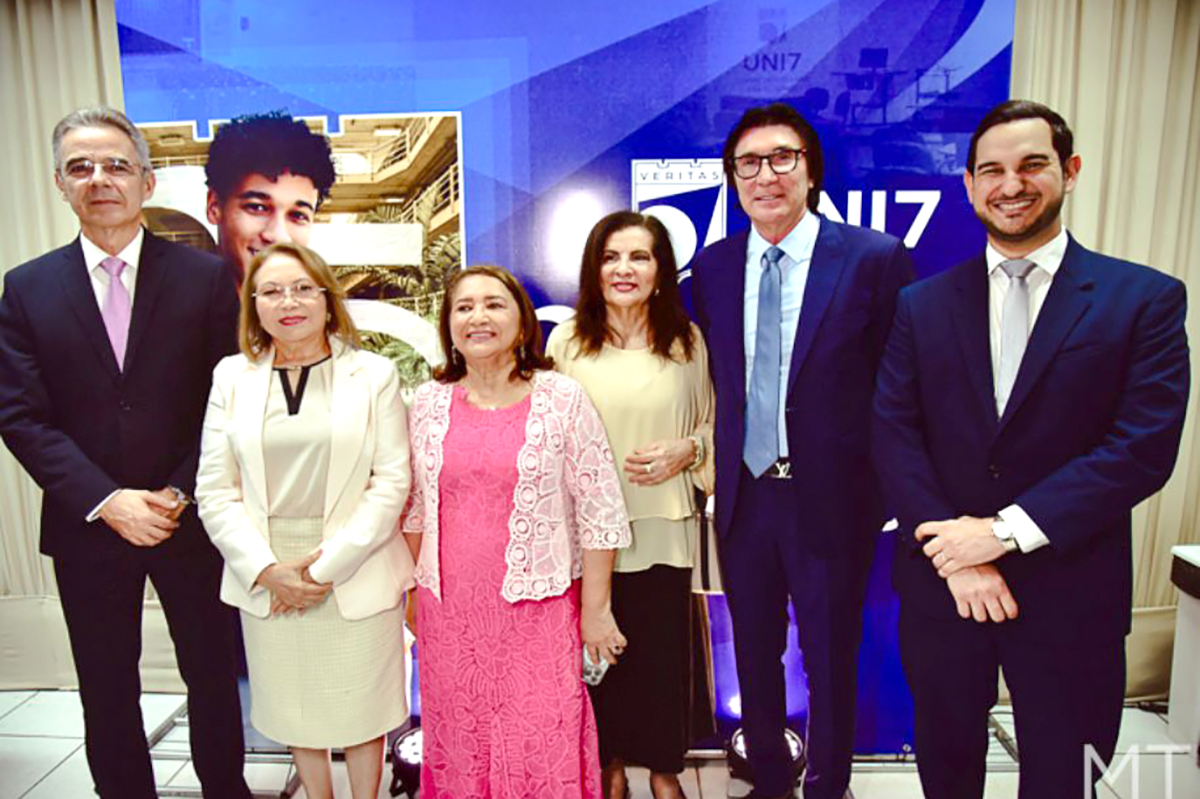Seis pessoas posam para uma foto oficial em frente a um painel azul da UNI7. Da esquerda para a direita: dois homens em ternos escuros, uma mulher de tailleur bege, uma mulher com vestido rosa rendado, a homenageada de blusa bege e, por fim, outro homem de terno e óculos.