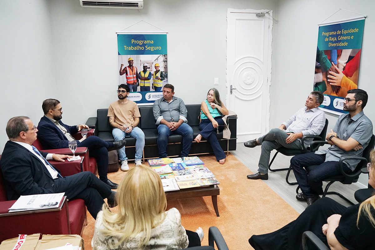 Em uma sala de reuniões com tapete claro, um grupo de pessoas está sentado em poltronas e sofás dispostos em semicírculo ao redor de uma mesa de centro repleta de cartilhas coloridas. Nas paredes ao fundo, destacam-se dois banners institucionais: "Programa Trabalho Seguro" e "Programa de Equidade de Raça, Gênero e Diversidade".